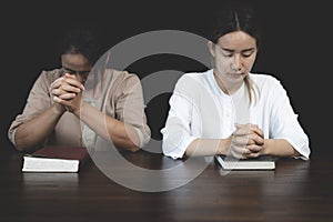 Two Christian women sit together and pray on a wooden table with blurred open Bible pages in their homeroom. Prayer for brothers,