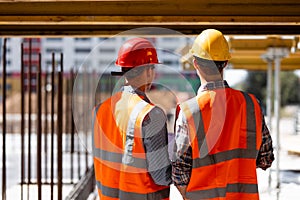 Two civil engineers dressed in orange work vests and helmets discuss the construction process on the building site near