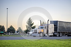 Two big rig classic semi trucks with turned on headlights with loaded semi trailers standing on the parking lot for rest take a