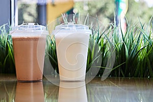 Two clear plastic glasses with lids filled with chocolate and vanilla milkshake on table. Blank mockup, background cafe