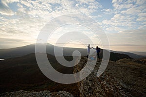 Two climbers standing on summit above clouds in the mountains holding hands. Silhouettes of hikers celebrating ascent on