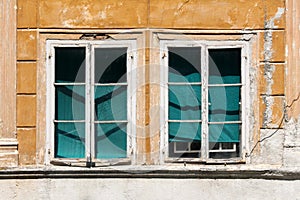 Two closed dilapidated windows with white wooden frames secured with strong metal bars and hidden with closed curtains