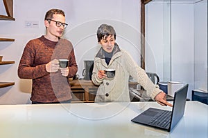 Two co-workers having a conversation while they consult a laptop and have coffee on a break from work in the office. Workaholics