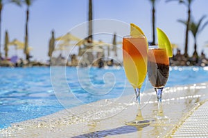 Two cocktails with natural juice and ice at the edge of the swimming pool. Orange cocktail with an orange slice and red