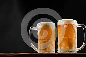 Two cold mugs with beer, with overflowing foam, on wooden table and dark background, space for writing