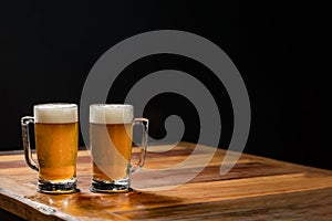 Two cold mugs with beer, with overflowing foam, on wooden table and dark background, space for writing