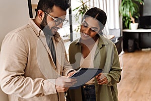 Two colleagues discussing business details and using tablet while standing in office space