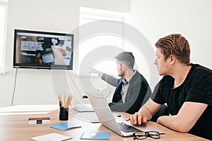 Two colleagues are sitting at table in the office. They are looking at TV-screan. They wear black clothes