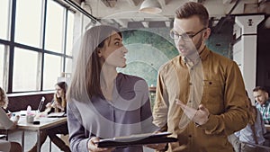 Two colleagues walking through the modern office and talking. Caucasian man and asian woman discussing the documents.