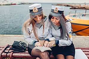 Two college women students of Marine academy reading book by sea wearing uniform. Friends studying