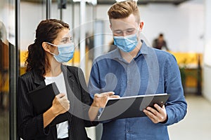 Two collegues discuss work and business wearing protective sterile medical masks standing in the corridor of the office.
