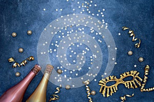 Two colorful champagne bottles with carnival mask, confetti stars and party streamers on blue background. Flat lay of christmas