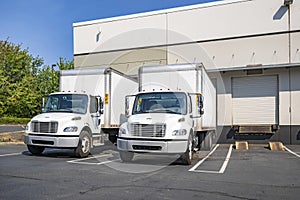 Two compact middle day cab rig semi trucks with box trailers standing on the warehouse parking lot with dock and gates loading