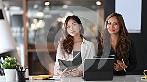 Two businesswomen sitting together in office and smiling to camera.