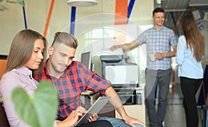 Two confident young people looking at touchpad while their colleagues working in the background.