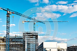 Two construction tower cranes at a construction site against a blue sky with clouds. Modern high-rise office building under