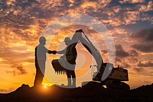 Two construction workers stand by an excavator at sunset