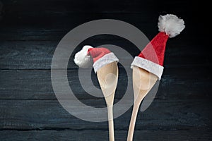 Two cooking spoons with red Santa Claus hats against a dark blue wooden background, Christmas invitation for a dinner or greeting