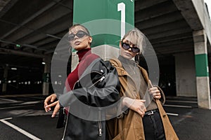 Two cool fashion sisters women in urban clothes with a leather jacket and a sweater wearing trendy sun glasses posing in the