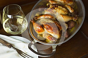 Two Cornish hens in an aluminum baking pan with handles on a wooden table, shown with a glass of white wine.