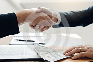 Two corporate businessmen shaking hands while one man places money on document in office room with corruption concept