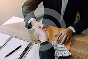 Two corporate businessmen shaking hands while one man places money on document in office room with corruption concept