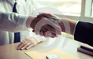 Two corporate businessmen shaking hands while one man places money on document in office room with corruption concept