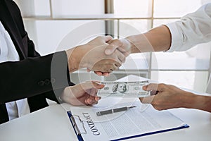 Two corporate businessmen shaking hands while one man places money on document in office room with corruption concept