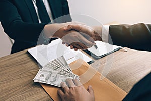 Two corporate businessmen shaking hands while one man places money on envelope document in office room with corruption concept