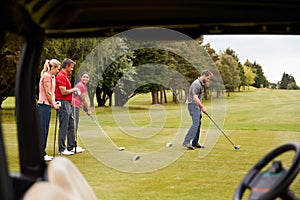 Two Couples Golfing Hitting Tee Shot Along Fairway With Driver With Buggy In Foreground