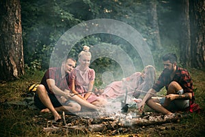 Two couples having picnic in woods. Bearded man and his best friend cooking sausages over fire. Young hikers around