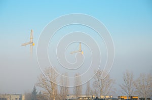 Two cranes in fog above rooftops and trees.