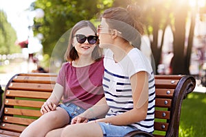 Two creative girls talk and laugh while sitting on bench outdoors. Young and funloving friends share ideas, thoughts and plan new