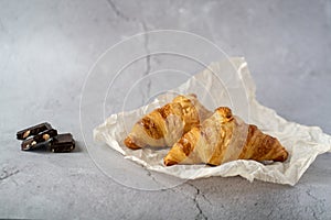 Two croissants lie on the table on a gray background next to three pieces of chocolate
