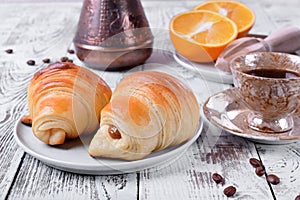 Two croissants of yeast dough on ceramic plate, freshly brewed coffee in a cup and oranges