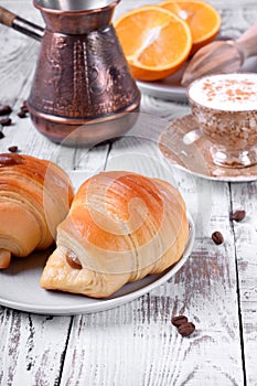 Two croissants of yeast dough on ceramic plate, freshly brewed coffee in a cup and oranges