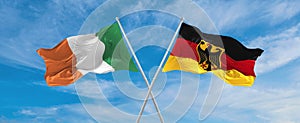 two crossed flags Germany and Ireland waving in wind at cloudy sky. Concept of relationship, dialog, travelling between two