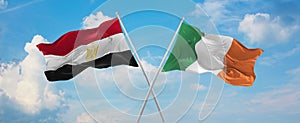 two crossed flags Ireland and Egypt waving in wind at cloudy sky. Concept of relationship, dialog, travelling between two