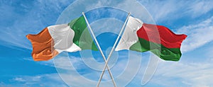 two crossed flags Madagascar and Ireland waving in wind at cloudy sky. Concept of relationship, dialog, travelling between two