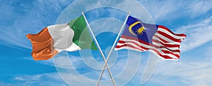 two crossed flags Malaysia and Ireland waving in wind at cloudy sky. Concept of relationship, dialog, travelling between two