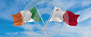 two crossed flags Malta and Ireland waving in wind at cloudy sky. Concept of relationship, dialog, travelling between two