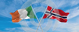 two crossed flags Norway and Ireland waving in wind at cloudy sky. Concept of relationship, dialog, travelling between two