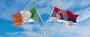 two crossed flags Serbia and Ireland waving in wind at cloudy sky. Concept of relationship, dialog, travelling between two
