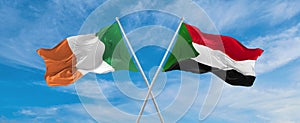 two crossed flags Sudan and Ireland waving in wind at cloudy sky. Concept of relationship, dialog, travelling between two