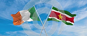 two crossed flags Suriname and Ireland waving in wind at cloudy sky. Concept of relationship, dialog, travelling between two