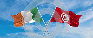 two crossed flags Tunisia and Ireland waving in wind at cloudy sky. Concept of relationship, dialog, travelling between two