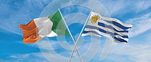 two crossed flags Uruguay and Ireland waving in wind at cloudy sky. Concept of relationship, dialog, travelling between two