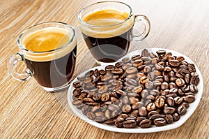 Two cups with black coffee, roasted coffee beans in saucer in heart shape on wooden table