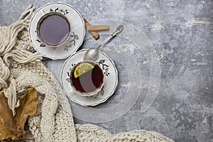 Two cups with black tea and lemon and saucers, lemon, cinnamon sticks, spoon, oak leaves and knitted scarf near, at gray backgroun