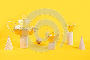 Two cups of camomile tea, transparent teapot on wood geometric shape, yellow background. Creative concept Natural